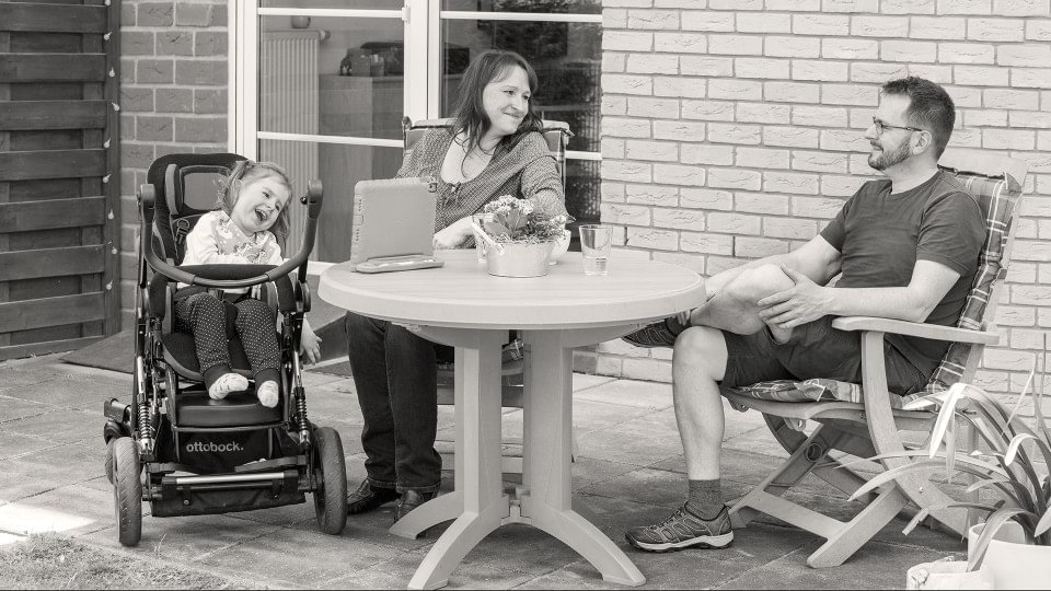 Girl with cerebral palsy sits with her parents and the Kimba Neo rehab buggy in the garden. 