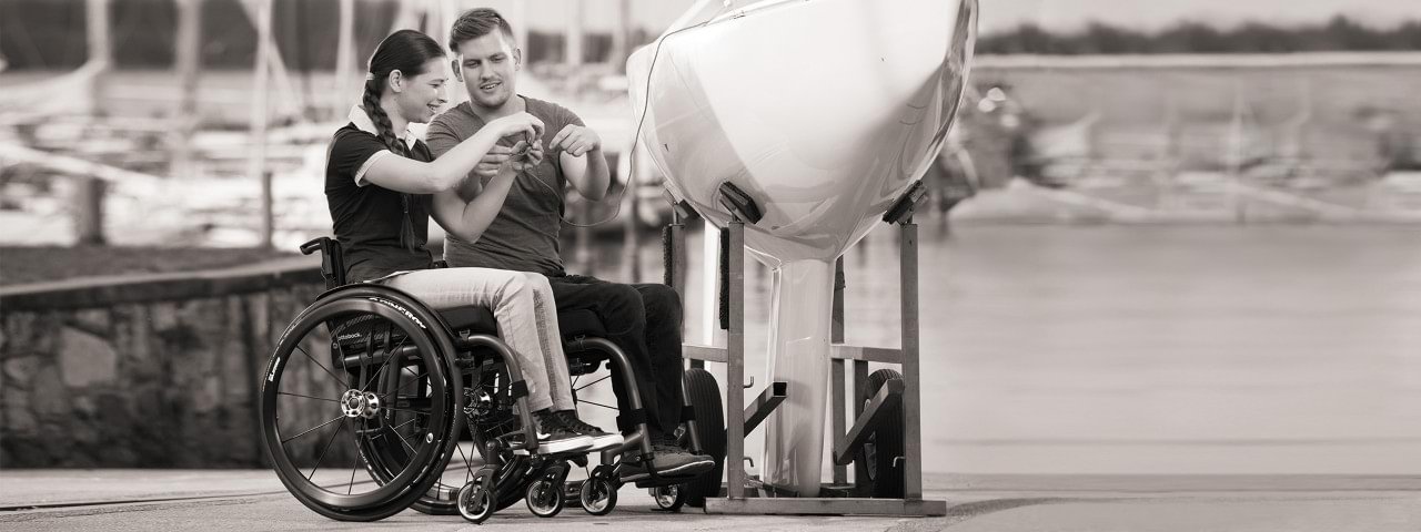 Zwei Rollstuhlfahrer sitzen an einem Hafen vor einem Boot.