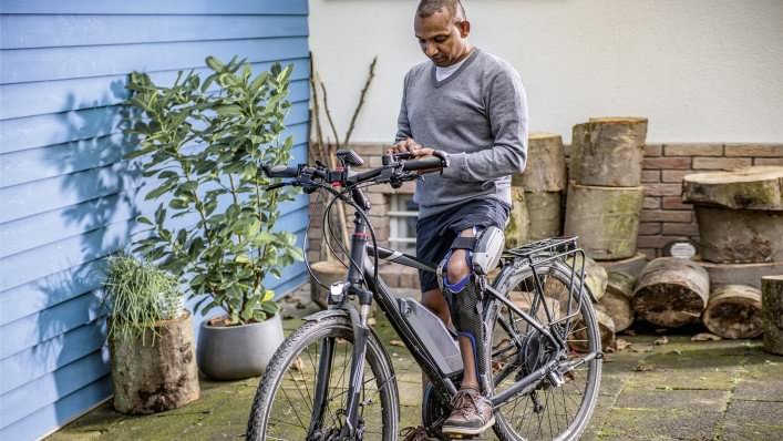 Melvin sits on his bicycle. He is wearing his computer-controlled C-Brace® orthotronic mobility system. He is holding his mobile phone, which shows the adjustment app on the display