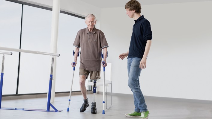 An amputee walks with two walking aids in the presence of his technician.