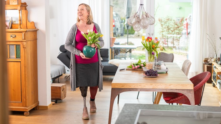 Anita wears her Trias prosthetic foot as she walks through the living room holding a flower vase in her hands. 