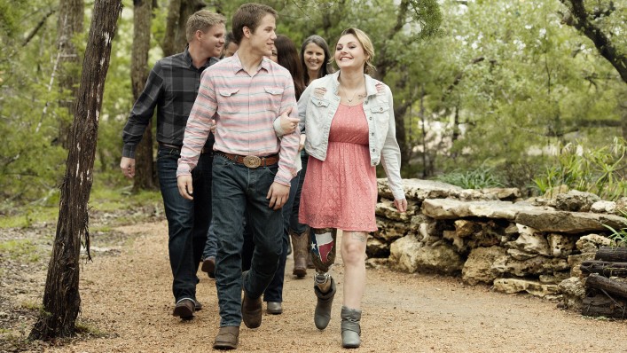 Three couples walking through the forest hand in hand behind one another. The woman in the first couple is wearing a C-Leg.