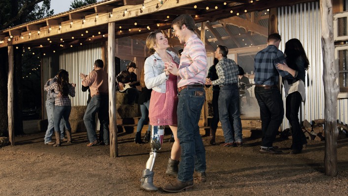 : A couple laughs and dances; she is wearing a C-Leg. A band and people dancing can be seen in the background.