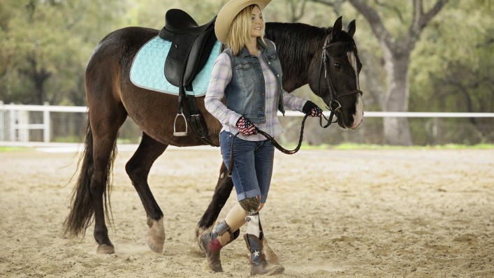 A cowgirl leading her horse across the riding ground. She wears a C-Leg on her right leg.