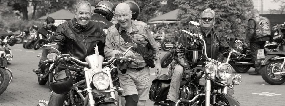 Georg steht völlig entspannt zwischen zwei Harleys und posiert lachend mit den Bikern für die Kamera.