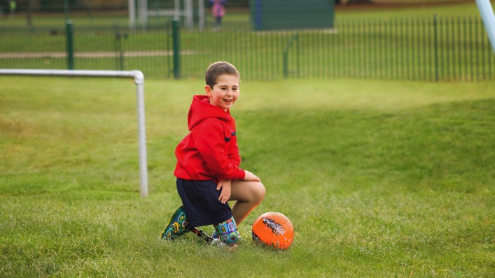 A small boy kneels on his prosthetic leg in the grass. A ball lies in front of him.